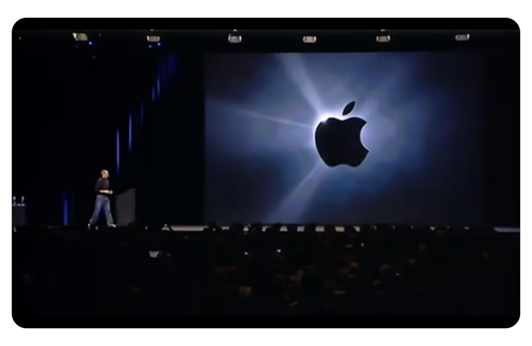 一个演讲场景，舞台上有一位穿着休闲装的男性站在左侧，背景是一个巨大的屏幕，屏幕上显示着发光的苹果公司标志，周围有光束效果，营造出科技感和庄重氛围。