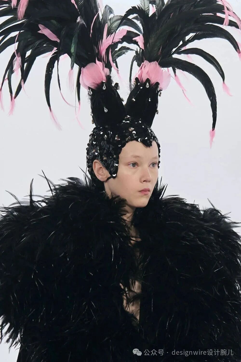 一位模特身着黑色羽毛主题时装，头戴高耸的黑色羽毛头饰，头饰顶部有大量黑色长羽与粉色短羽交错装饰，中央点缀两团粉色绒球及垂挂水钻；头饰下方为覆盖头顶的黑色亮片与珠饰发箍，造型如兔耳或角状；模特肩部及上身被浓密蓬松的黑色羽毛披肩覆盖，整体风格华丽、前卫且具戏剧性，背景为纯白色，右下角有水印文字。