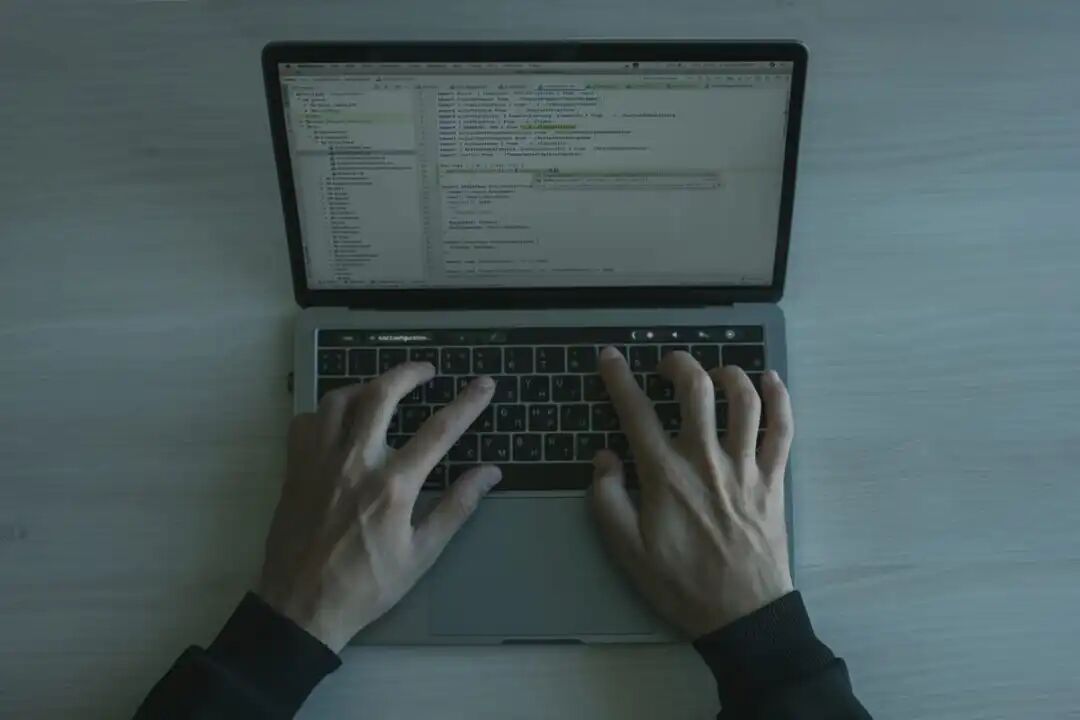 俯视视角下，一双手在 MacBook 键盘上敲击，屏幕上显示着代码编辑器界面