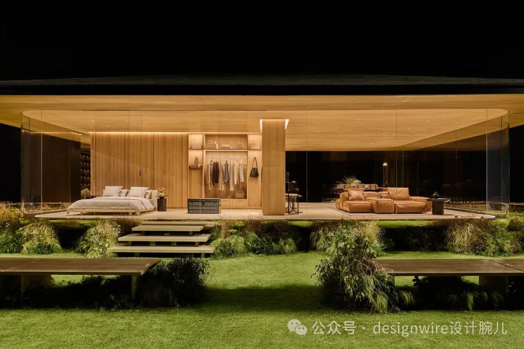 夜晚拍摄的现代风格住宅展示场景，建筑主体为玻璃幕墙结构，内部以暖色木质饰面为主，分隔出卧室与客厅区域。左侧卧室配有浅色床品的双人床、床头柜及背景墙储物系统；中间区域设衣帽间，挂有衣物并陈列包袋；右侧为棕色皮质组合沙发区，搭配小桌与落地灯。建筑外为草坪景观，设有木质台阶与长凳，整体灯光柔和，营造温馨静谧氛围。