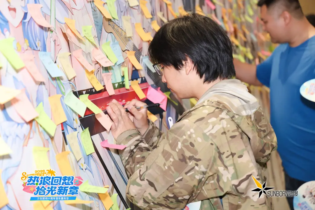 一名指挥官在少女前线十周年嘉年华留言墙前，用笔在粉色便签上写下留言