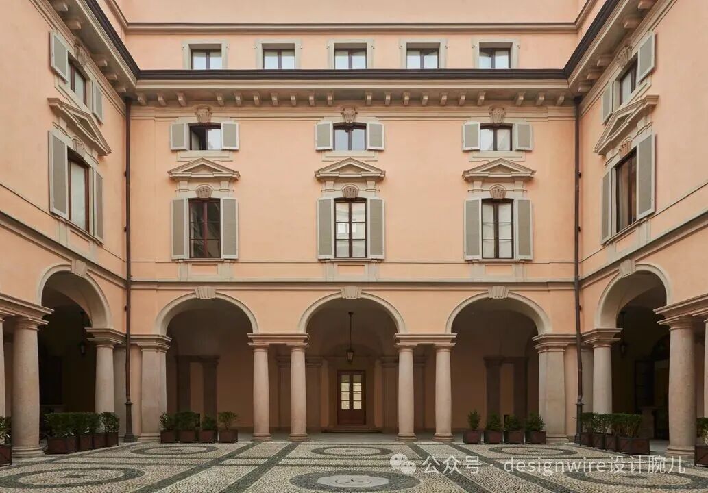 一座欧洲古典风格的建筑庭院，四面围合，外墙为浅粉橙色，窗户配有灰绿色百叶窗，窗框上方有三角形山花装饰；底层为连续拱廊，由多根圆柱支撑，拱廊内可见一扇木门；地面为黑白相间的几何图案鹅卵石铺地，拱廊两侧摆放着成排绿植盆栽；画面右下角有水印文字“公众号 · designwire设计腕儿”