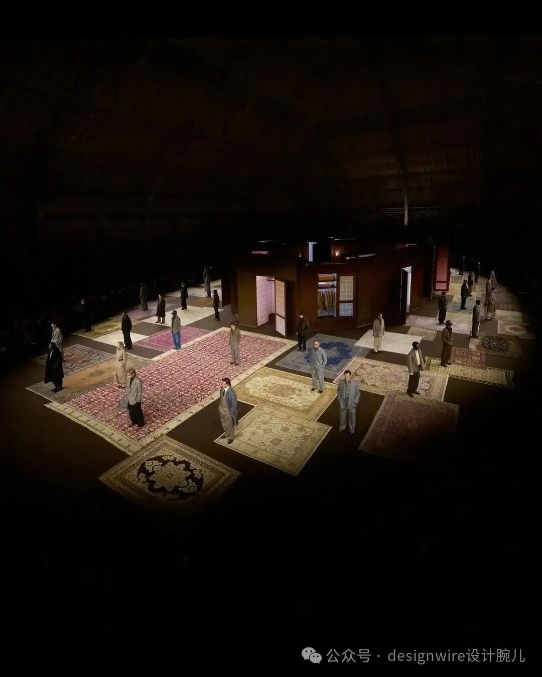 高角度俯拍的室内时尚秀场场景，整体环境昏暗，地面铺满多块大小不一、图案各异的地毯，包括红色花纹、米色古典纹样、蓝色几何图案等；多位模特或观众站立于地毯之上，穿着不同风格的服装，以灰、蓝、黑为主色调；中央区域设有一座深棕色木质结构小屋，设有门和窗，内部可见衣架与衣物；画面右下角有水印文字“公众号 · designwire设计腕儿”；灯光集中照射在地毯与人物区域，形成明暗对比强烈的舞台效果。