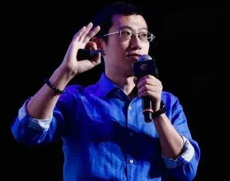 一名戴眼镜男子手持麦克风发言，身穿蓝色衬衫，背景为黑色
