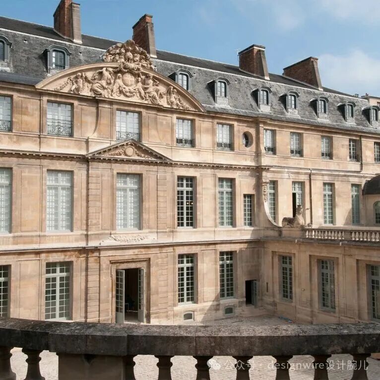一座典型的法国古典主义风格建筑，米黄色石材立面，多层结构，窗户为白色窗框格子窗，屋顶为深灰色瓦片，可见多个老虎窗、烟囱及带铁艺护栏的窗户；中央上方有大型浮雕装饰，包含人物与徽章元素；底层中央设双开木门，部分窗户开启。前景可见石质栏杆，下方为庭院地面。右下角有水印文字“公众号 designwire设计腕儿”。