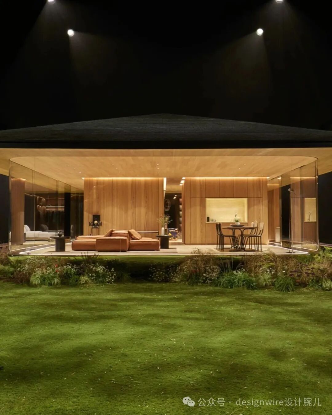 夜晚拍摄的现代风格住宅外观，建筑采用大面积玻璃幕墙与木质饰面，内部暖色灯光透出。室内可见橙棕色皮质沙发组合、圆形餐桌配四把椅子，背景为木质墙面及嵌入式灯带照明。屋顶呈平缓坡度，上方有三束聚光灯照射建筑顶部。前景是修剪整齐的绿色草坪，草坪边缘有低矮灌木和野花植被。右下角有水印文字。