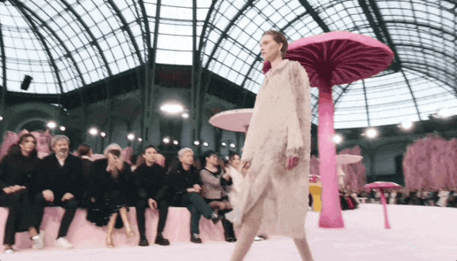 一位模特在室内大型玻璃穹顶建筑内走秀，身穿米白色长款毛绒质感长裙，正从画面右侧向左侧行走。秀场布置充满梦幻感，设有多个巨大粉色蘑菇造型装饰物，伞盖为粉红色，菌柄为亮粉色；观众席沿T台两侧设置，坐满穿着深色服装的观众，部分观众正在拍照。背景可见复杂的金属桁架结构和顶部照明灯。画面右下角有'CHANEL'水印。