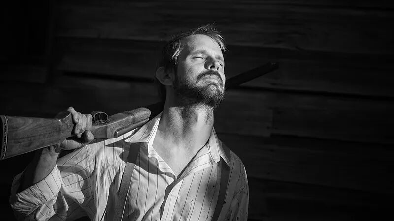 一位闭目沉思的男子扛着猎枪，背景为木板墙