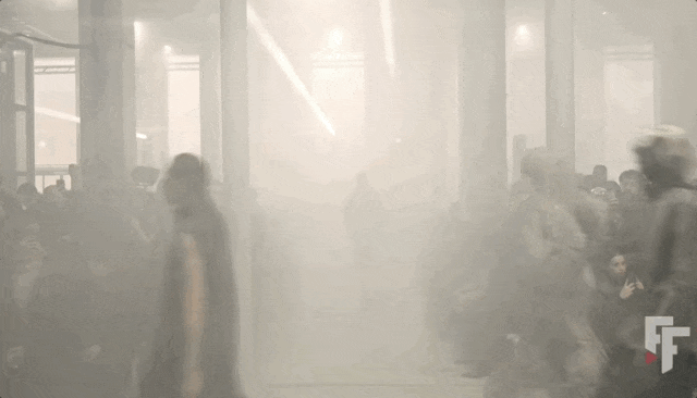 一张在烟雾弥漫的室内秀场拍摄的动态画面，呈现时装秀场景。画面中央是一条T台，两侧坐满观众，多根粗大立柱贯穿空间。多位模特依次从烟雾中走出，穿着深色系服装，因烟雾和运动模糊导致轮廓与细节不清，但可见其行走姿态。顶部有几道斜向白色光束穿透烟雾，营造戏剧性氛围。右下角有红白相间的'FF'字母水印（Fashion Forward或类似品牌标识）。整体色调为灰白单色，颗粒感明显，具有电影感与朦胧艺术效果。