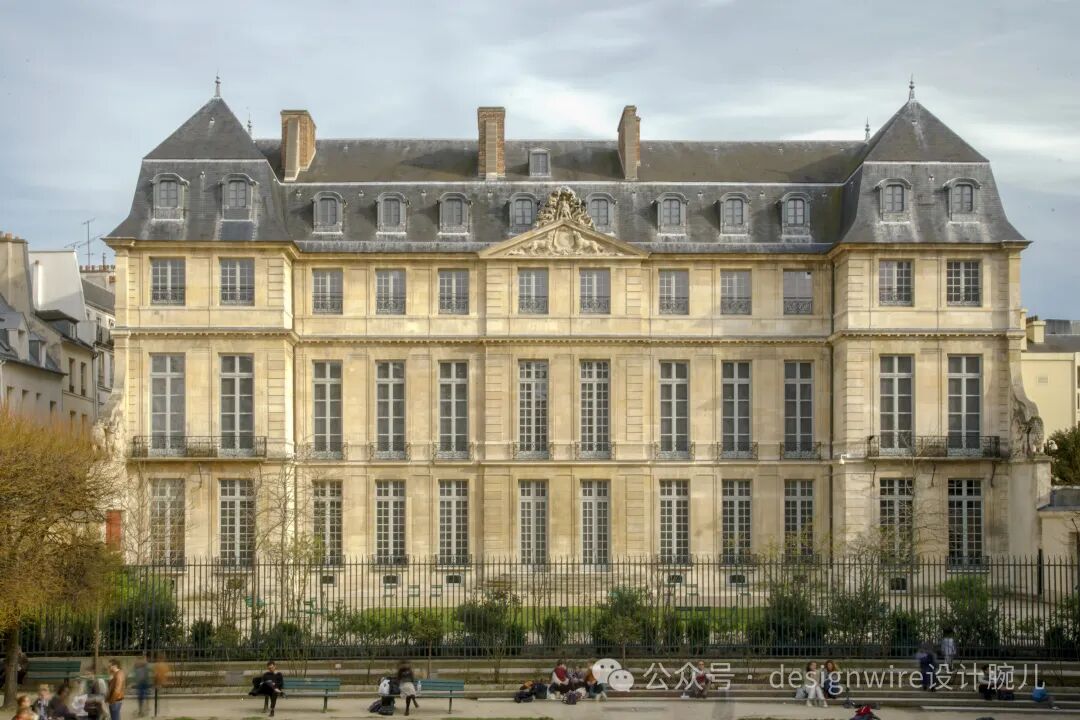 一座三层古典风格的米黄色石砌建筑，拥有对称布局和深灰色斜屋顶，屋顶设有多个老虎窗与烟囱；中央立面有装饰性山花浮雕，各层窗户为多格玻璃窗并配黑色铁艺栏杆；建筑前方是黑色金属围栏，围栏内有绿植，围栏外广场上有行人散步、坐卧休憩；画面右下角可见水印文字‘公众号 · designwire设计腕儿’；背景为多云天空及周边其他建筑。