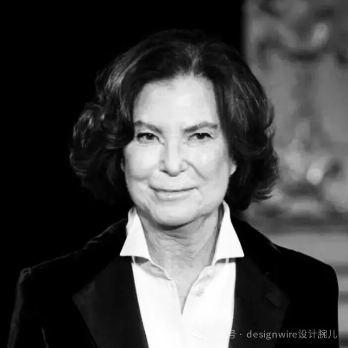 一张黑白肖像照，一位年长女性正面朝向镜头，面带温和微笑。她留着及肩的卷发，发色较深，发型蓬松自然。身穿白色翻领衬衫，外搭深色西装外套，衣领清晰可见。背景为纯黑色，右侧有模糊的浅色装饰物轮廓，整体构图为中近景特写，光线从正面打来，突出面部轮廓与表情细节。