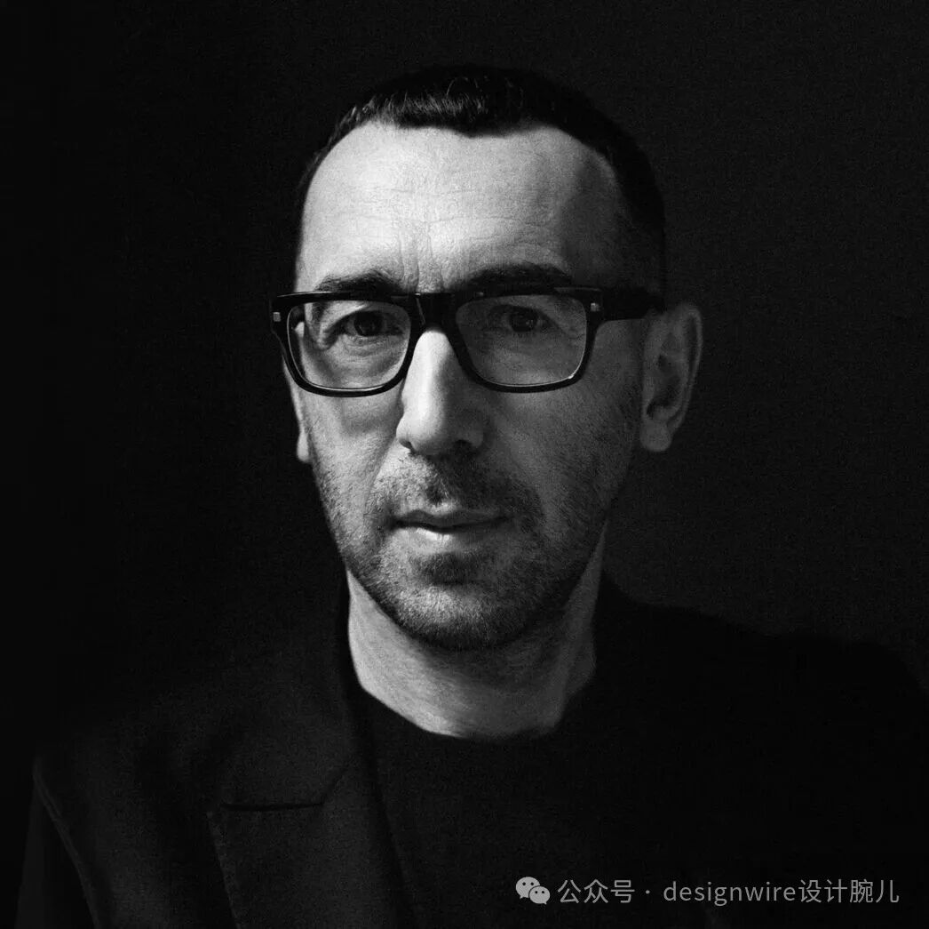 一张黑白风格的男性肖像特写照片，背景为纯黑色，人物位于画面中央偏右，面部正对镜头，表情平静。该男子戴一副黑色粗框眼镜，发型短而整齐，有轻微胡茬，穿着深色高领上衣和外套。画面光线从左上方打来，在其面部形成明暗对比，突出五官轮廓。右下角有白色水印文字：公众号 · designwire设计腕儿