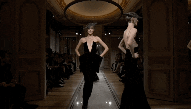 一场高级时装秀现场，模特们身着黑色礼服在室内T台走秀。T台两侧坐满观众，背景为古典风格的装饰墙面与大型圆形吊灯，整体光线偏暗，聚焦于T台中央。画面中可见多位模特依次走过：第一位模特穿深V领无袖黑色连体裤装，戴灰色羽毛帽；第二位模特穿露背黑色长裙，戴宽檐帽；第三位模特穿亮片点缀的深V露肩长裙，戴黑色小礼帽；第四位模特穿同款亮片长裙，正转身离开。所有服装均以黑色为主，设计优雅且富有戏剧性。