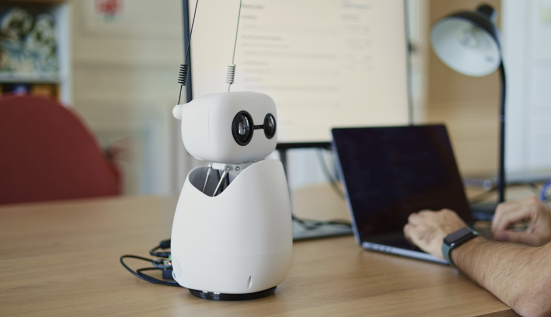 一个白色桌面机器人，拥有圆形头部和天线，背景有人操作电脑