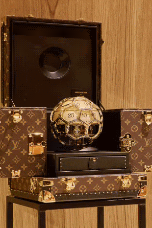 图片展示了一个奢华的路易威登（Louis Vuitton）限定款足球艺术品，被陈列在木质背景前的黑色金属高脚台上。主体是一个金色与白色相间的足球，表面有精致的浮雕图案和数字“25”、“30”，置于一个黑色底座之上；底座下方叠放着两个打开的路易威登经典老花硬箱，箱体为棕色配金色LV字母与花卉图案，配有金色锁扣与铆钉；上方大箱盖内侧为黑色绒面，中央有圆形凹陷设计；整体呈现高端定制、收藏级商品的展示效果。
