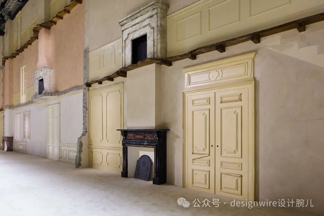 室内场景，呈现仿古建筑立面结构，墙面为米色与浅粉色，部分墙体有破损露出砖石结构。画面右侧有一对米黄色双开门，门上有古典浮雕线条装饰；门前设黑色大理石壁炉，壁炉上方有红木装饰，壁炉前地面放置一块深色弧形炉挡。左侧可见另一组米黄色镶板门、一扇白色窗框窗户及一个石砌壁炉开口。顶部有木质横梁悬挑，整体为舞台布景或展览装置风格，右下角有水印文字。