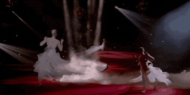 舞台表演场景，深色背景下红色圆形舞台中央有白色烟雾效果，多名舞者身着飘逸白色长裙在舞台上进行舞蹈动作，部分舞者被吊索悬吊于空中，灯光从上方投射形成光束，观众席环绕舞台可见，整体氛围梦幻且富有戏剧性。