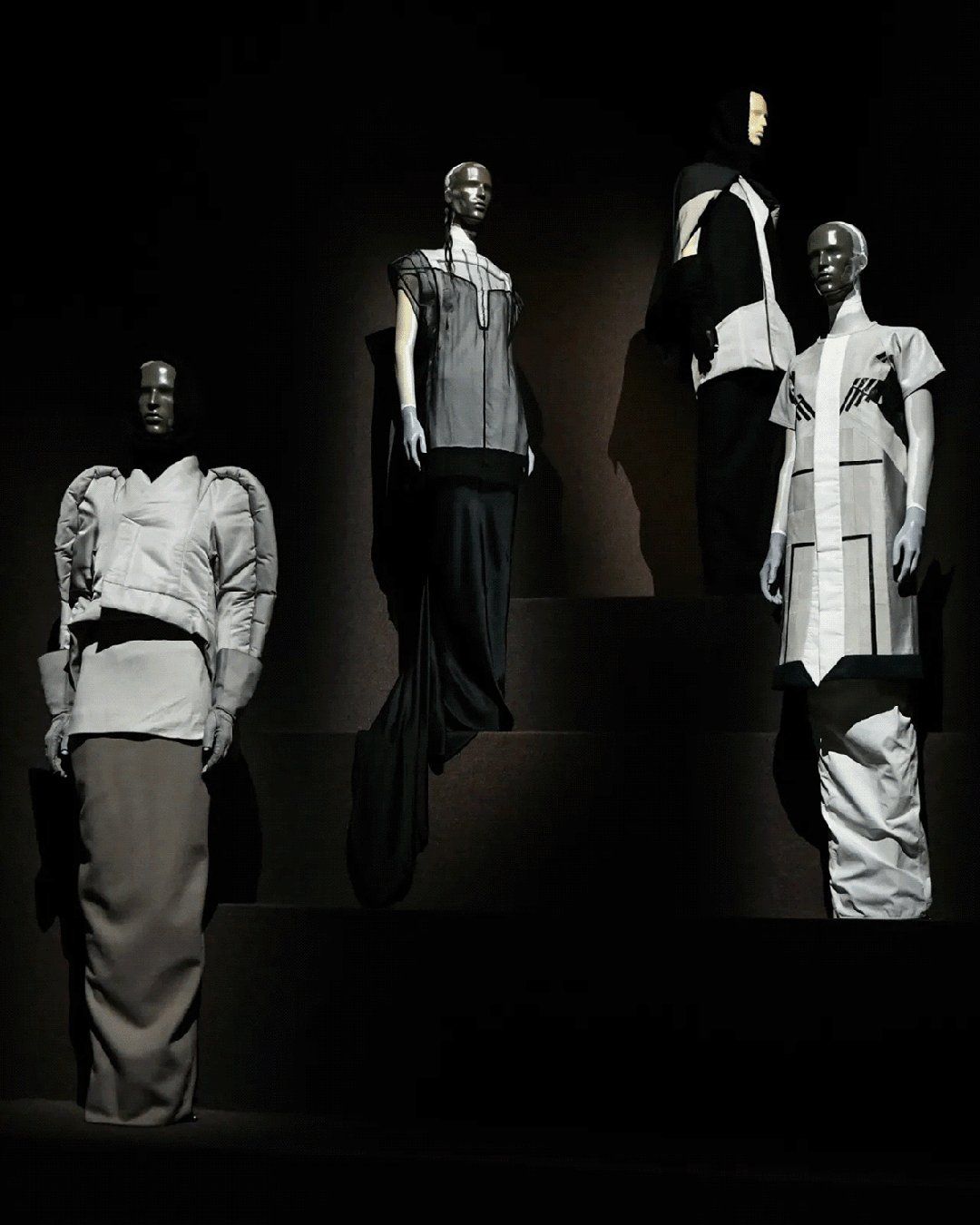 四张图片展示了一场高级时装展览的多个场景，均为黑白或暖色调灯光下的展厅布景。每张图中均有多具无表情、金属质感或哑光肤色的模特假人，站立于阶梯式展台上，穿着风格前卫、结构复杂、强调几何线条与垂坠感的服装。第一张图为黑白色调，左侧模特穿蓬松短上衣配长裙，中间两模特分别着透明拼接长裙与黑白拼色连体装；第二张图为米白/奶油色系，中央有长拖尾礼服，背景挂有抽象画作；第三张图为棕褐色调，背景墙上挂有古典油画及小型雕塑画框，模特服饰多为贴身剪裁与褶皱设计；第四张图为透过深色幕布窥视的视角，两位模特身着棕色套装，下方有说明牌。整体氛围庄重、艺术性强，突出服装的建筑感与叙事性。