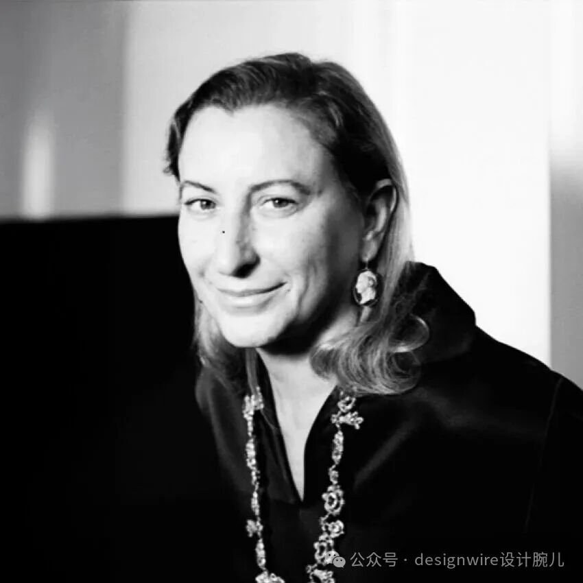 一张黑白肖像照，一位中年女性侧身面向镜头，面带微笑，目光温和。她留着及肩波浪卷发，发色较浅，左侧脸颊有一颗小痣。身穿深色高领或立领上衣，颈部佩戴一条由多个装饰性吊坠组成的长项链，耳垂上戴着一枚复古风格的圆形雕花耳环。背景为简洁的明暗对比墙面，左侧为深色区域，右侧为亮白色墙面，光线从右前方打来，在面部形成柔和阴影。图片右下角有水印文字。