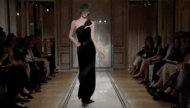 一位模特在室内时装秀T台上走秀，身穿黑色单肩长裙，裙身饰有亮片，腰间系有宽腰带。她头戴一顶巨大、装饰繁复的黑色宽檐帽，帽面布满闪亮珠饰并带有网状遮面设计，遮住了眼部。模特姿态优雅，一手叉腰，正沿铺有浅色条纹的T台向前行走，两侧坐满观众。背景为古典风格的室内空间，墙面有雕花装饰，顶部有射灯照明，整体色调偏暗，突出服装与配饰的光泽感。