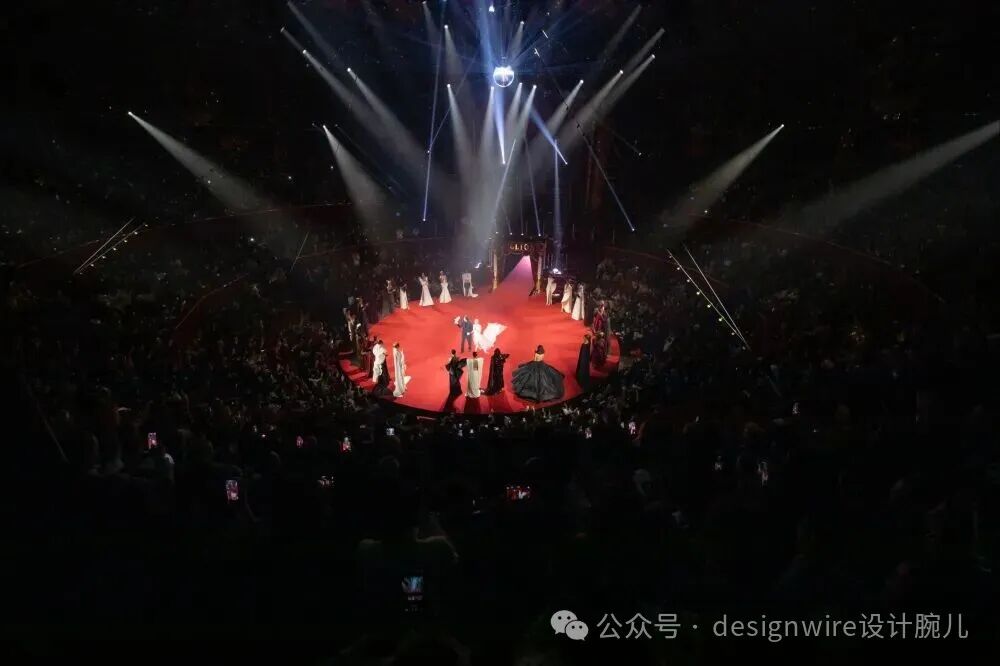 高角度俯拍的大型室内演出场景，中央为圆形红色舞台，多名身着礼服的表演者围成半圆站立，其中一位穿着黑色蓬蓬裙礼服的女性位于前方中央，另有几位穿白色或深色服装的表演者在舞台中央互动；舞台上方多束白色聚光灯从不同角度照射，形成明亮光柱；观众席环绕舞台，黑暗中可见大量观众及举起的手机闪光灯；右下角有水印文字‘公众号 · designwire设计腕儿’