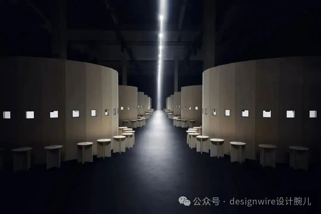 一个昏暗的室内空间，中央有一道垂直的强光束从天花板射下，照亮地面形成光柱。两侧对称排列着多组米色弧形隔间结构，每个隔间墙面上嵌有多个方形发光小窗。隔间前方摆放着多张圆形矮桌和配套的无靠背圆凳，整体布局呈纵深走廊状，营造出静谧、未来感与极简主义氛围。右下角有水印文字。