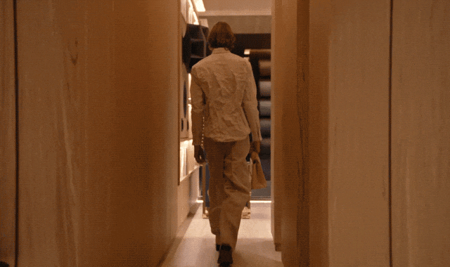 一名金发人士背对镜头，身穿白色褶皱长袖衬衫与浅色宽松长裤，右手提着一个米色手袋，正沿着一条两侧为木质墙面的狭长走廊向前行走。走廊尽头可见深色置物架及部分家具轮廓。随后画面切换至夜晚的现代风格住宅外部全景：建筑采用大面积玻璃幕墙与木质立面，内部灯光温暖明亮，可清晰看到客厅区域的棕色沙发、餐桌椅以及另一名在厨房区域活动的人物；该名穿白衬衫者正从右侧走向餐桌区域，整体环境呈现极简、温馨且富有设计感的居住空间氛围。