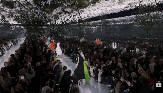一场Dior时装秀现场，T台两侧坐满观众，中央为白色反光T台。天花板悬挂着大量白色花朵与绿色枝叶，营造出樱花林或花园的沉浸式布景。多位模特正沿T台走秀，前景一位模特身着黑色长裙搭配亮绿色披肩，其后方可见穿白色及橙色礼服的模特。画面右下角有Dior品牌标识及YouTube播放按钮图标。