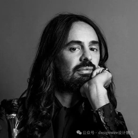 一张黑白肖像照，一位长发、留有浓密胡须的男性，他右手托腮，手指上戴有戒指，目光直视镜头，神情沉思。他身穿带有复杂花纹装饰的深色外套，背景为纯灰色渐变，光线从左侧打来，在面部形成明暗对比。右下角有水印文字“公众号 · designwire设计腕儿”，字体较小，呈白色。