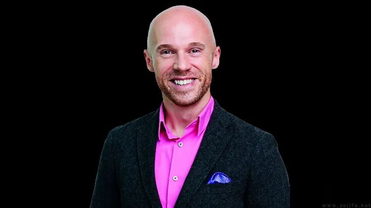 Robert Hallock 面带微笑，身穿亮粉色衬衫和深灰色西装，在黑色背景下接受访谈