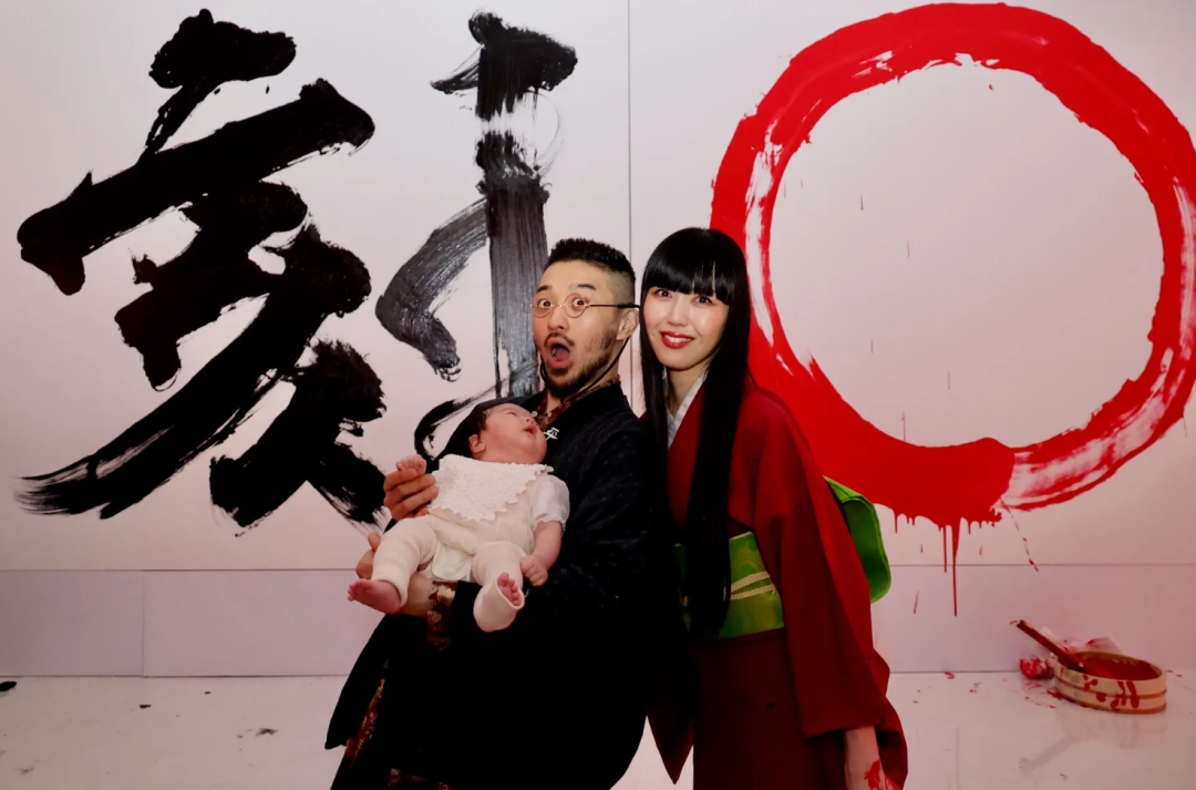 一张家庭合影，背景为白色墙面，左侧有黑色大号毛笔书法“武”字，右侧有红色圆形墨迹，下方地面散落墨点；画面中央一名戴眼镜、留短发胡须的男性穿深色和服，抱着一个穿白色连体衣的婴儿，表情夸张张嘴；其右侧一名黑长直发、齐刘海的女性穿红色和服配绿色腰带，面带微笑；右下角地面上有一个盛有红墨汁的砚台及毛笔，墨汁有滴落痕迹。