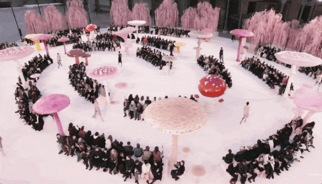 俯瞰视角下的时尚秀场场景，地面为白色，中央区域布置有多个大型蘑菇造型装置，伞盖呈粉色、红色及浅色斑点纹理，菌柄为粉红色；观众席呈环形围绕中央秀道，人群密集就坐，多为深色正装；秀场背景是成排的粉色垂柳状装饰树；若干模特身着浅色服装在场地中走动，整体色调以粉白为主，营造梦幻童话氛围。