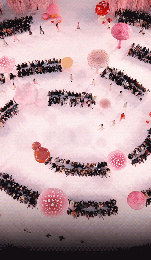 一场以梦幻童话风格为主题的时装秀现场，场地铺满白色地面，布置有多个巨型蘑菇造型装置（粉白相间、红白相间），背景是垂落的粉色柳枝状装饰。观众席环绕T台呈环形或半环形分布，坐满穿着正式的观众。多位模特身着设计前卫的服装在T台上走秀，服装风格多样，包括黑色亮片套装、红裙配白色羽毛外套、黑白印花长裙等；部分模特佩戴夸张头饰或披风。现场有摄影师在场拍摄，整体氛围浪漫、奇幻、高级。