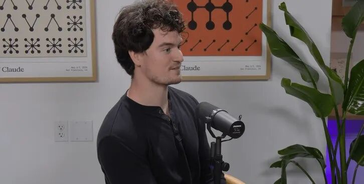 卷发男性身穿黑色亨利领上衣，侧坐于麦克风前，背景挂有Claude主题装饰画