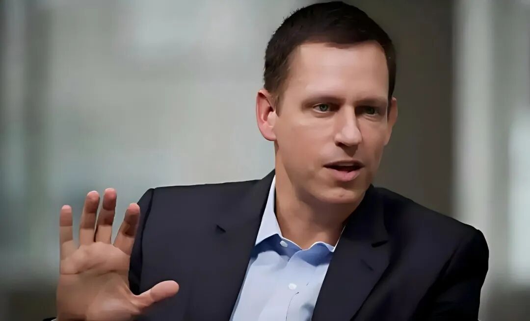 彼得·蒂尔身着深色西装，侧面站立，左手抬起做手势，背景为模糊的室内环境