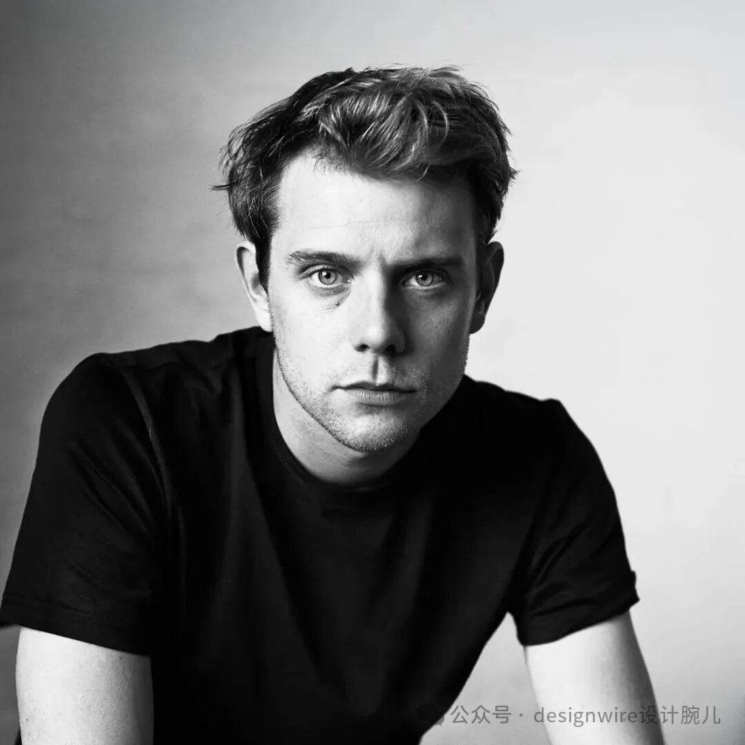 一位黑白肖像照，一名年轻男性正视镜头，头发蓬松略显凌乱，眼神深邃，面部有少量胡茬，身穿黑色短袖T恤，背景为纯浅灰色渐变，整体光线柔和，人物位于画面中央偏左，构图简洁突出面部表情。右下角有水印文字。