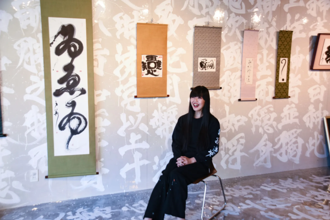 一位留着黑色长发、戴墨镜的女性坐在金属框架椅子上，面带笑容。背景为浅色墙面，布满白色书法笔触的涂鸦式文字；墙上悬挂多幅卷轴作品：左侧为绿色底、白纸黑墨大字草书（可见‘為’‘而’等字），中间自左至右依次为米黄底小方块篆刻风格作品（含‘思’‘心’字样）、灰底带点纹样卷轴（内有‘歸’字）、浅粉底竖幅（含小字落款）、深绿底带斜纹卷轴（内有问号‘？’），最右侧可见部分装裱画框（含‘書’字）。地面为灰色反光材质，映出墙面上文字倒影。
