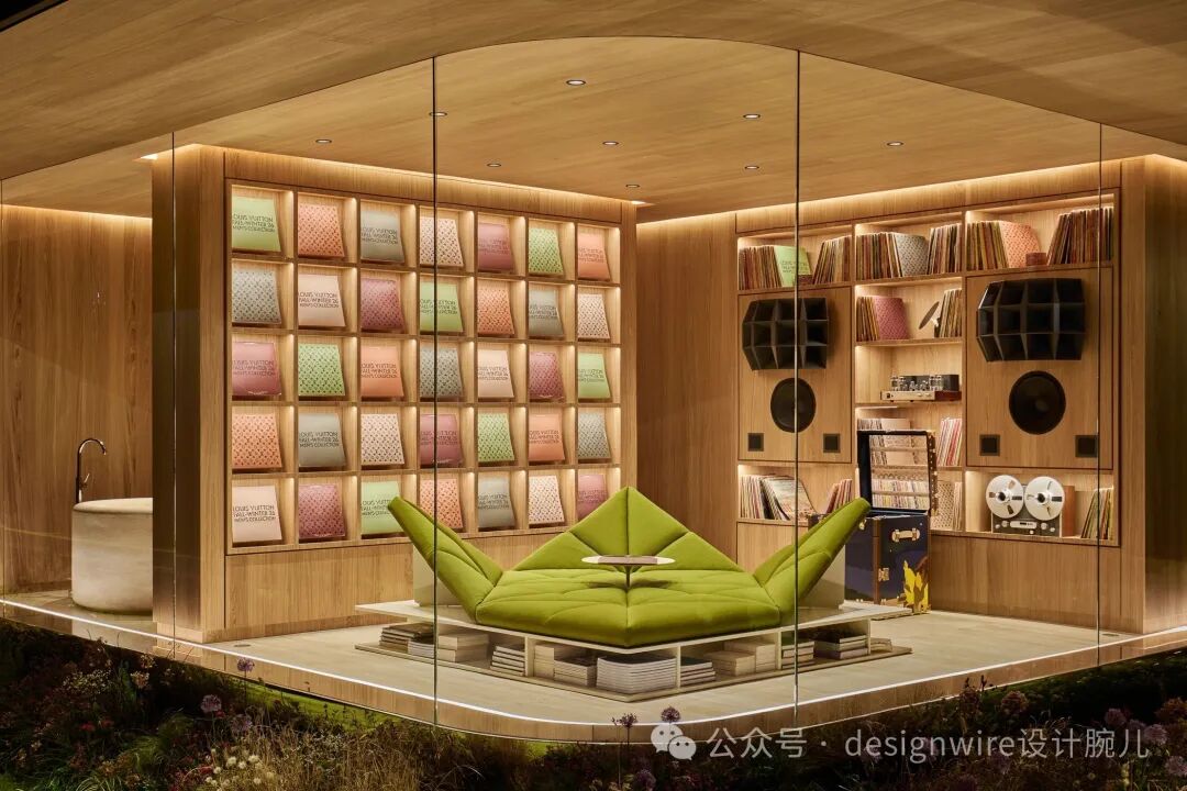 一个木质主题的沉浸式展示空间，被玻璃围合，内部设有弧形木质墙面与嵌入式照明。左侧墙面上排列着多格彩色封面的书籍或册子，颜色包括粉、绿、棕等；右侧为书架组合，陈列着大量书籍、黑胶唱片、复古音响设备（含圆形低音单元）、一台老式磁带录音机及一个蓝色行李箱。中央区域设有一张亮绿色模块化软垫沙发，呈对称展开造型，下方由白色层板与书籍堆叠支撑，前方配小圆桌。左下角可见一个白色独立浴缸与金属水龙头。整体空间以暖木色为主调，顶部有嵌入式射灯，底部边缘有灯带照明，前景为模拟自然植被的景观。右下角有水印文字“公众号 · designwire设计腕儿”。