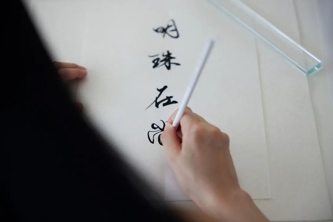 一只手握着白色毛笔正在白纸上书写黑色书法，已写有三个竖排汉字‘明珠在’，第四个字‘夜’正被书写中；画面左上角可见另一只手扶着纸张，右上角放置一个透明长条形压尺，整体为近景特写，光线柔和，背景简洁。