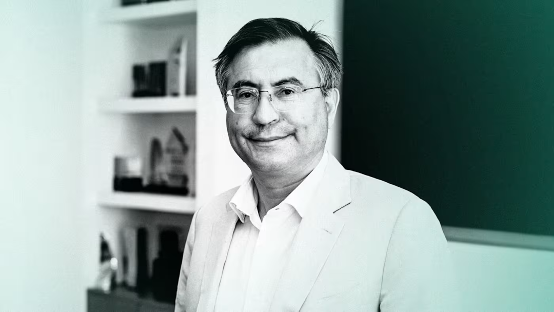 面带微笑的戴眼镜中年男性,身穿浅色西装,站在室内场景中