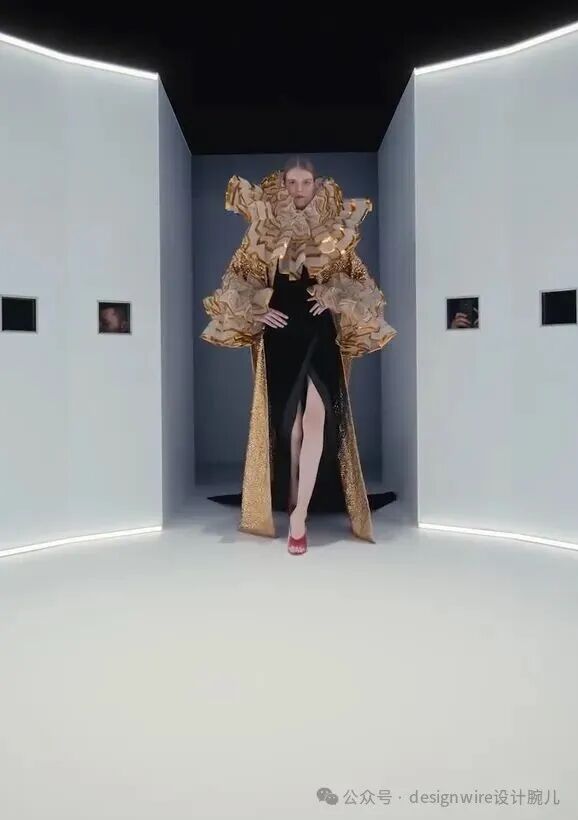 一位女性模特站在白色现代展厅中央，身穿黑色高开衩长裙搭配金色立体层叠装饰的夸张袖部与肩部造型，裙摆两侧延伸出金色亮片长拖尾，脚穿红色高跟鞋。背景为纯白墙面与黑色天花板，墙边嵌有数个黑色方形画框，内含模糊人像；地面与天花板边缘有环形LED灯带照明。画面右下角有水印文字。