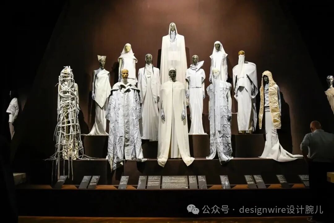 展厅内陈列着多件白色系服装设计作品，展示于阶梯式展台上，背景为深色。服装风格前卫、雕塑感强，材质多样，包括编织结构、层叠褶皱、几何切割等工艺；部分服饰覆盖全身并配以兜帽或头饰，造型类似宗教或仪式性长袍；中央位置有一件高挑的纯白长袍立于最高台，两侧对称分布其他款式；左侧可见一件由细绳/金属条缠绕构成的立体结构服装；右侧有金色装饰细节的长裙；前景底部设有多个黑色说明牌；右下角可见一名参观者背影，画面右下角有水印文字‘公众号 · designwire设计腕儿’。