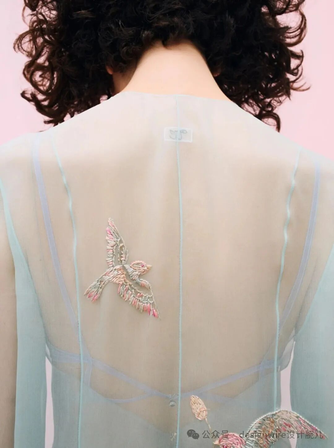 一位卷发女性背对镜头，穿着浅蓝色透明薄纱上衣，背部中央有精致的飞鸟刺绣图案，羽毛以粉、白、绿等多色丝线绣制，细节丰富；肩带与后背交叉绑带结构清晰可见；颈部下方贴有一枚白色小标签，印有模糊字母或符号；画面右下角有水印文字‘公众号 designwire设计腕儿’及部分彩色刺绣装饰边缘。