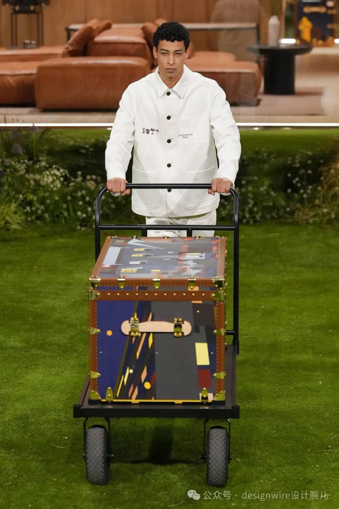 一名年轻男性模特身穿白色长袖工装外套与同色系长裤，站在绿色草坪上，双手推着一辆黑色金属手推车。手推车上放置一个大型复古风格行李箱，箱体为棕色皮革边角配金色铆钉与锁扣，箱面贴有抽象艺术图案，包含蓝色、深灰、黄色等色块及几何线条；背景为室内布景，可见棕色皮质沙发、小桌及绿植装饰，整体场景似时装秀T台。右下角有水印文字“公众号 · designwire设计腕儿”。